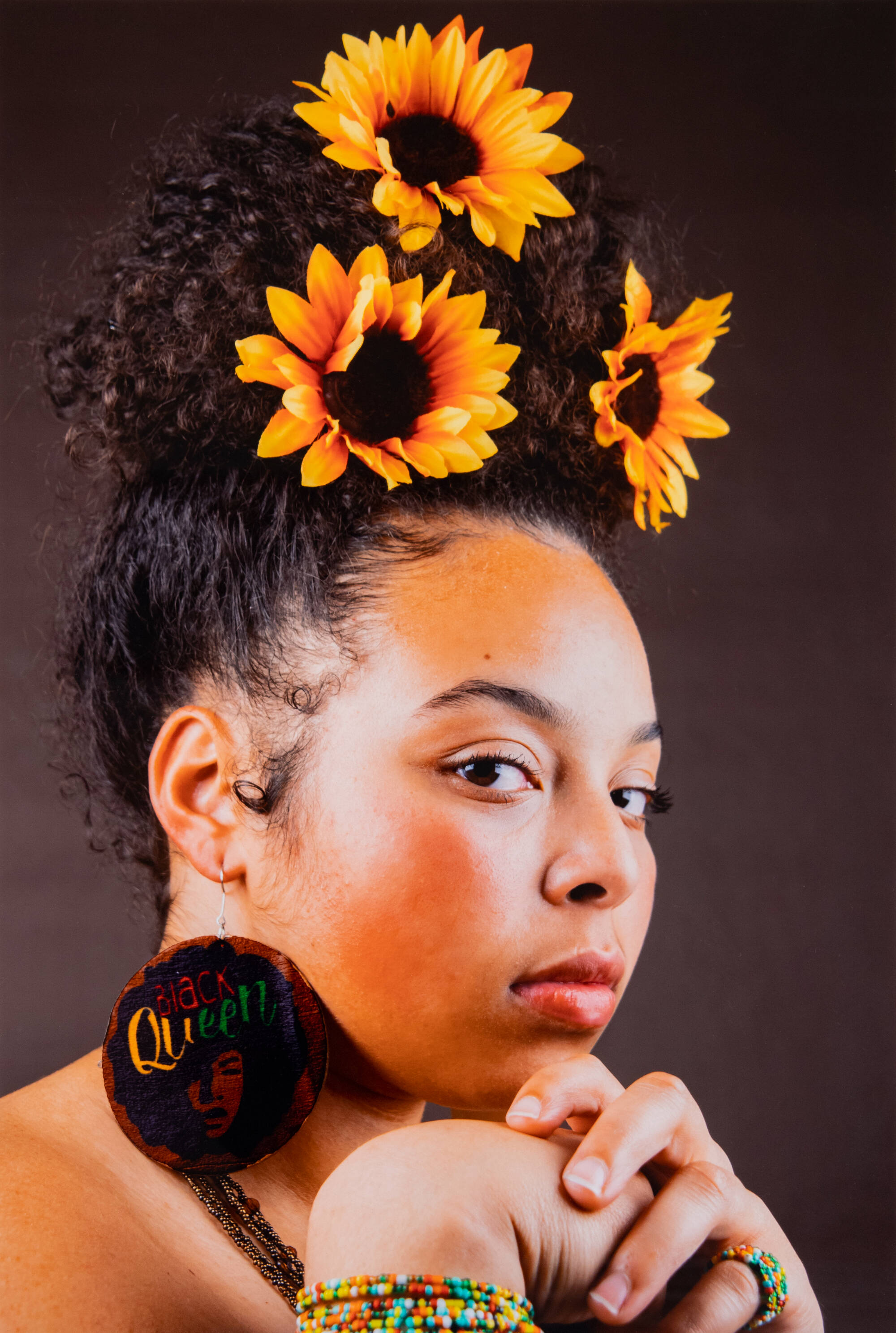 Color photograph of a side profile of a woman with sunflowers in her hair and large earrings that say "queen."